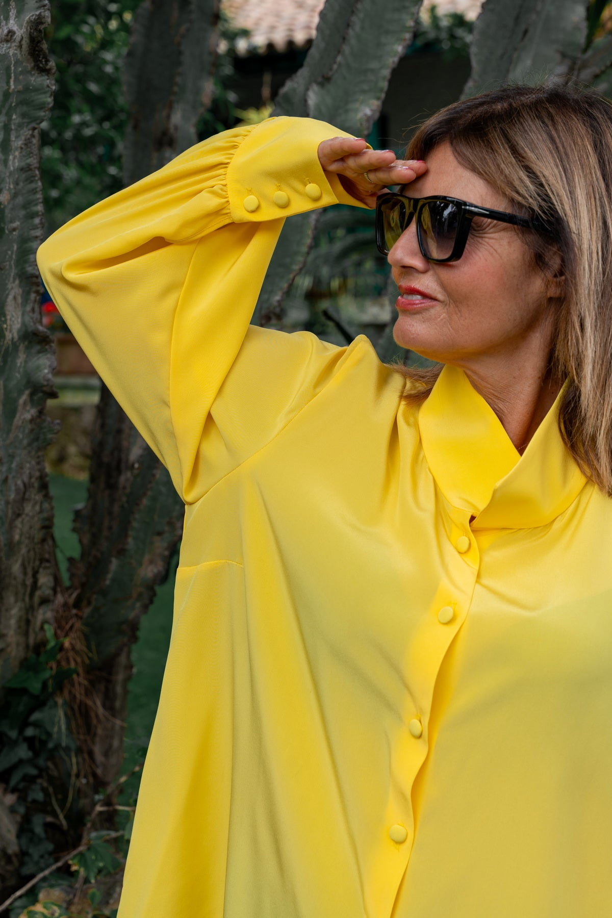 Camicia lunga seta crepe de chine gialla indossata da una donna elegante, con occhiali da sole e vista sul giardino.