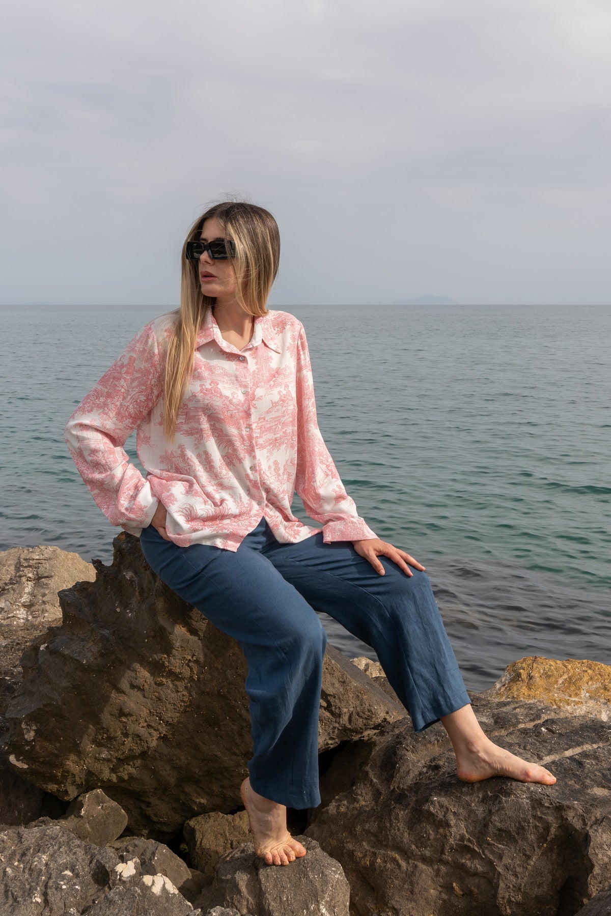 Donna che indossa la Camicia Elisabeth in Toile de Jouy rosa, seduta su rocce vicino al mare.