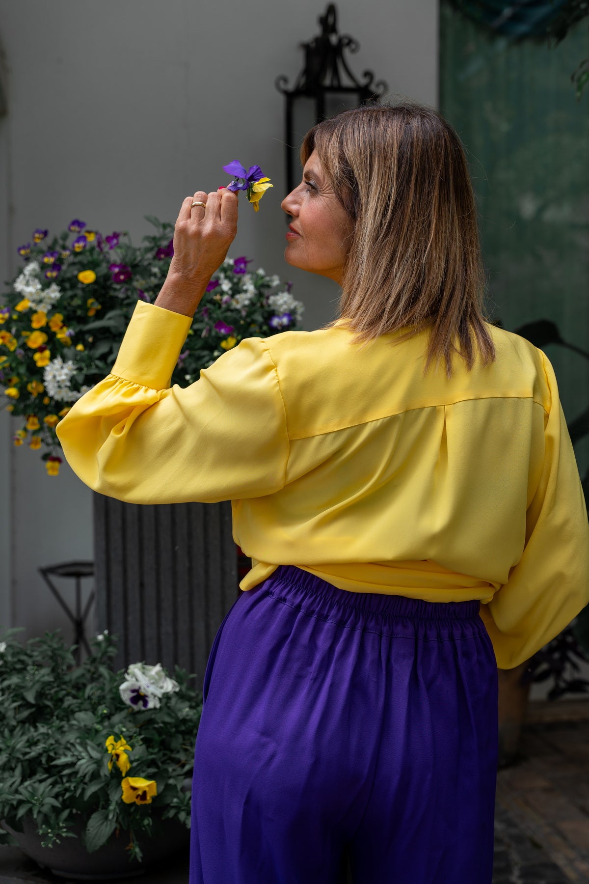 Donna che indossa una camicia gialla e pantaloni Marlene in viscosa viola, circondata da fiori colorati.