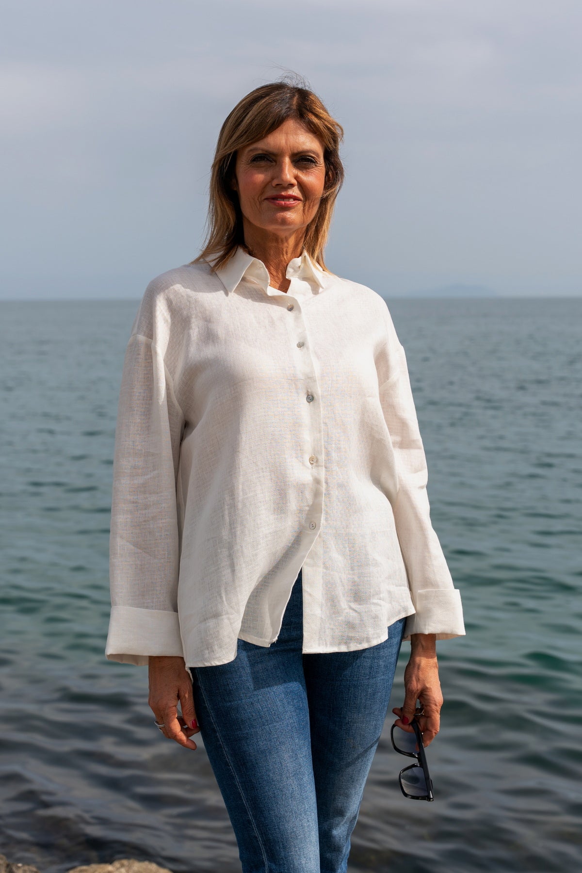 Camicia Capri in lino ricamata bianca indossata da una donna in riva al mare, estate26, lusso accessibile.
