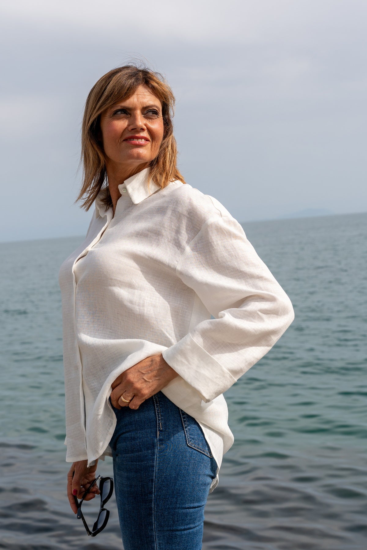 Camicia Capri in lino ricamata bianca indossata da donna con jeans vicino al mare.
