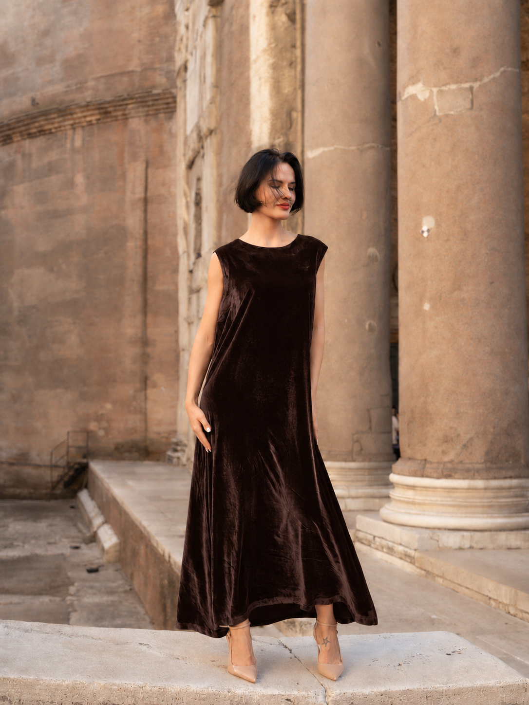 Abito lungo in velluto di seta marrone indossato da una donna elegante di fronte a colonne storiche.
