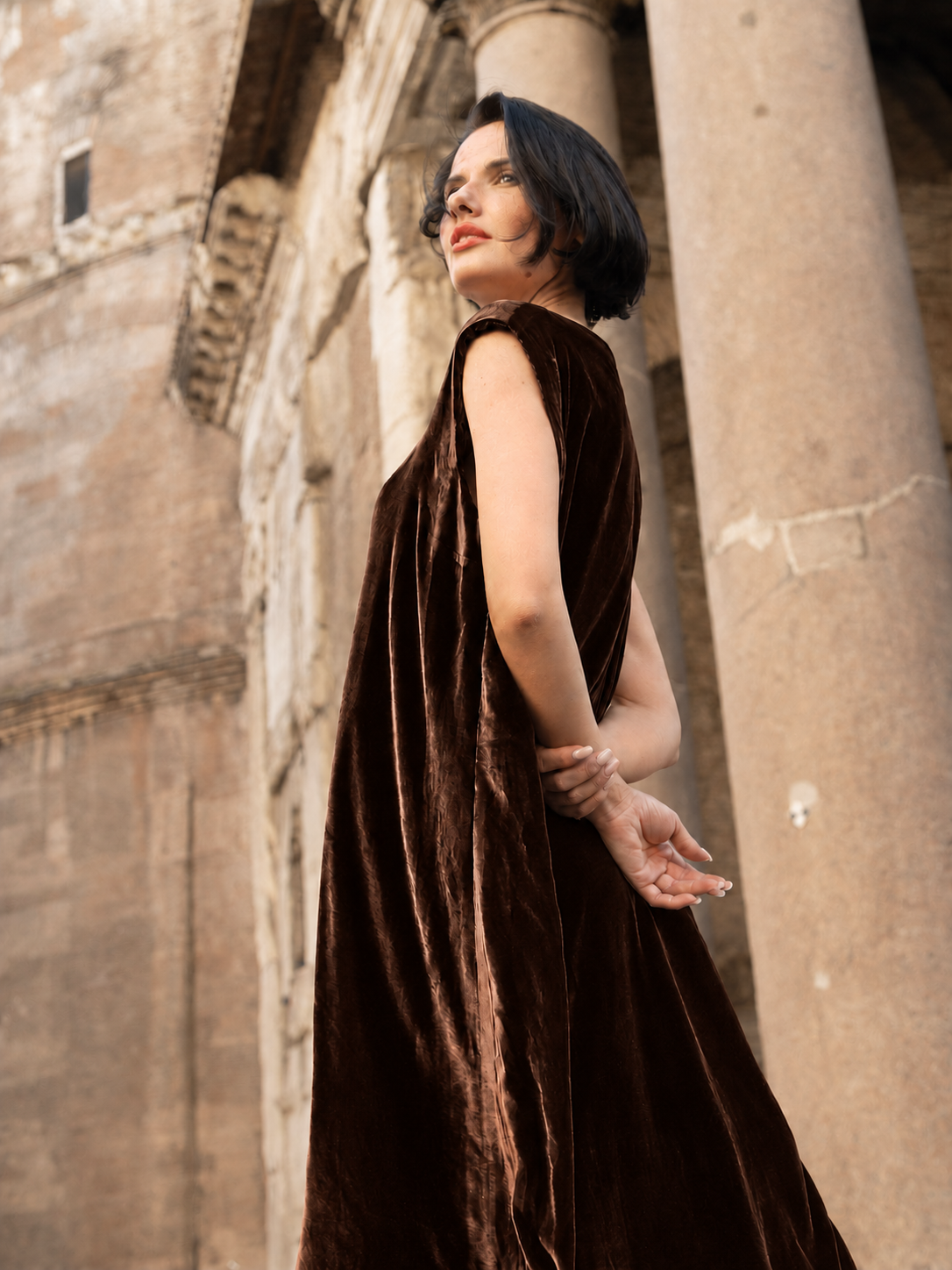Abito lungo in velluto di seta marrone indossato da una donna elegante davanti a colonne storiche.