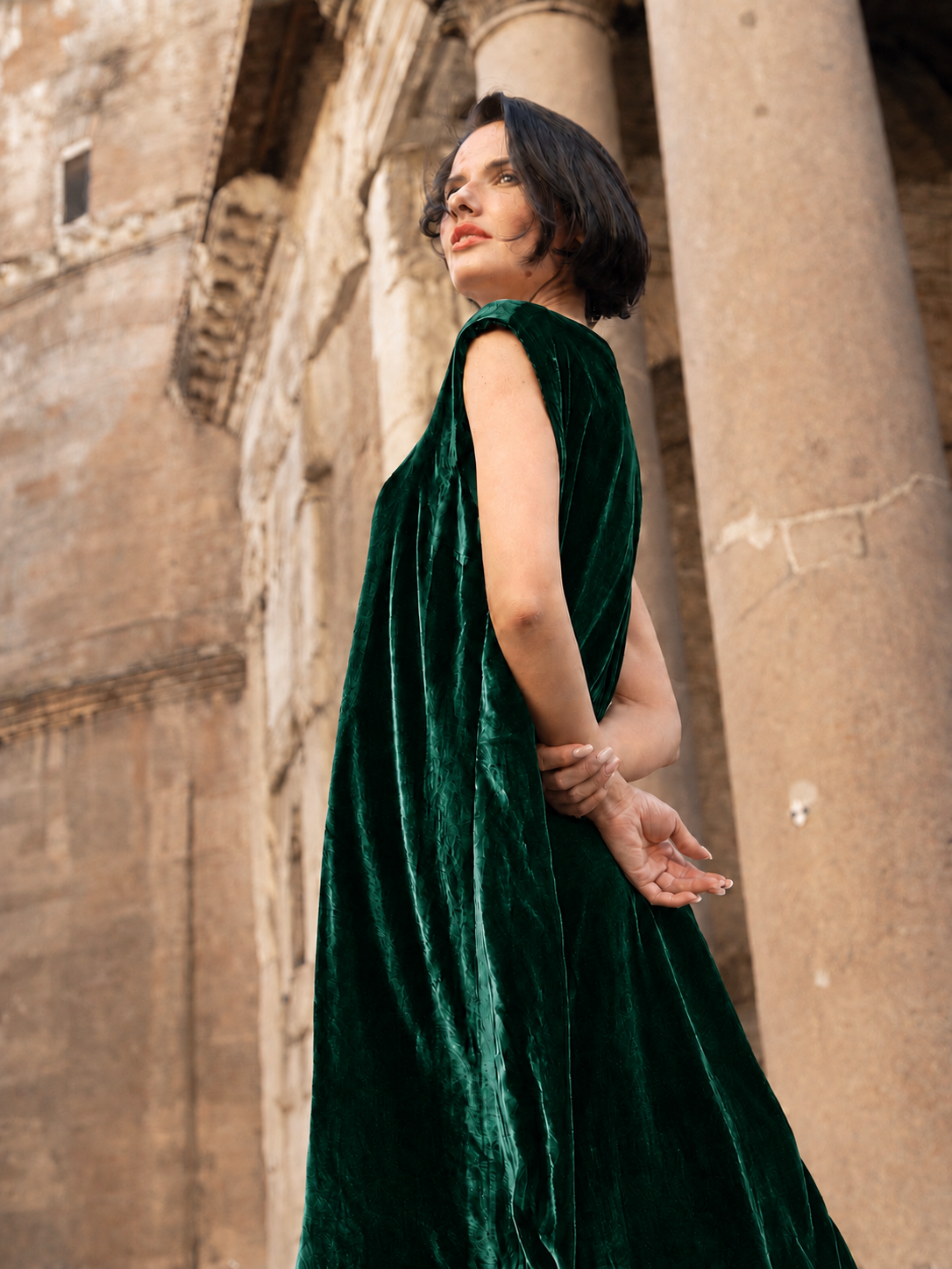 Abito lungo in velluto di seta verde indossato da una donna elegantemente posizionata davanti a colonne storiche.