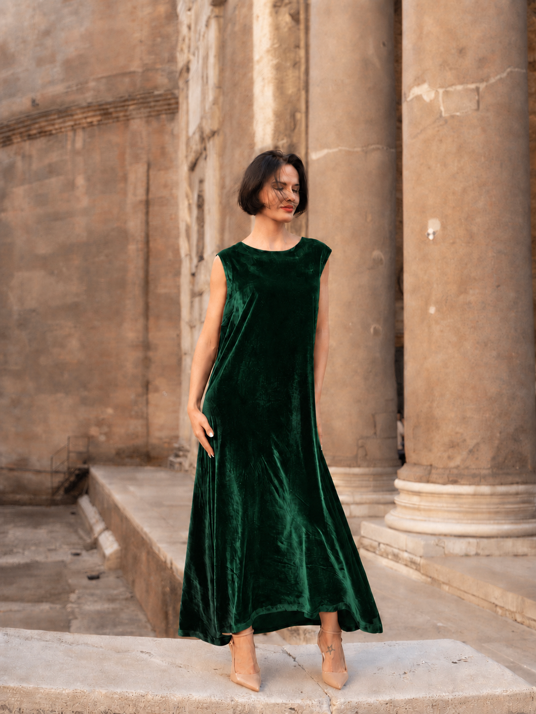 Abito lungo in velluto di seta verde indossato da una donna elegante in un contesto architettonico raffinato.