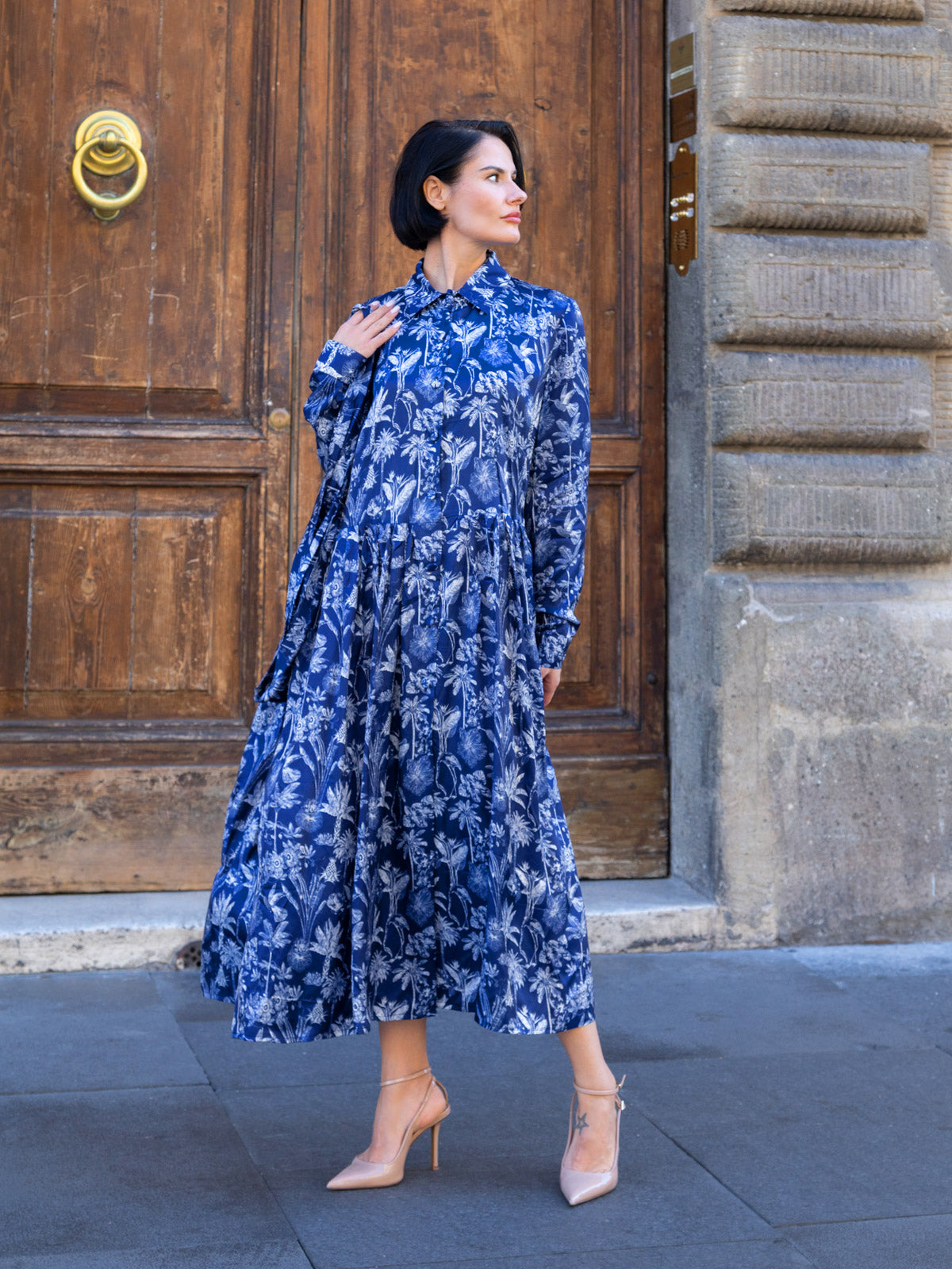 Abito lungo viscosa Liberty con stampa floreale blu e avorio, davanti a una porta in legno.