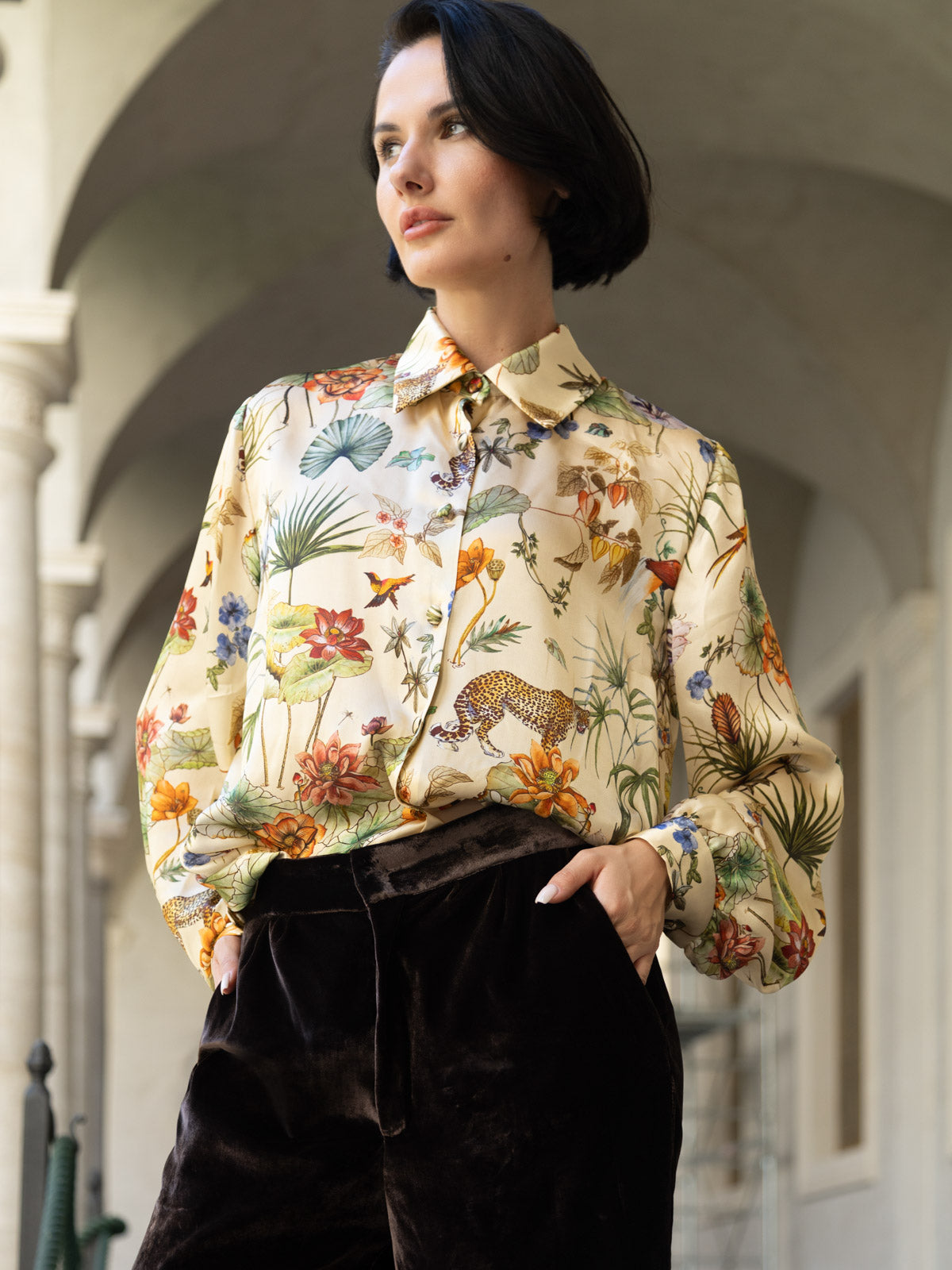 Camicia in seta avorio con stampa botanica indossata da una donna, abbinata a pantaloni di velluto scuro.
