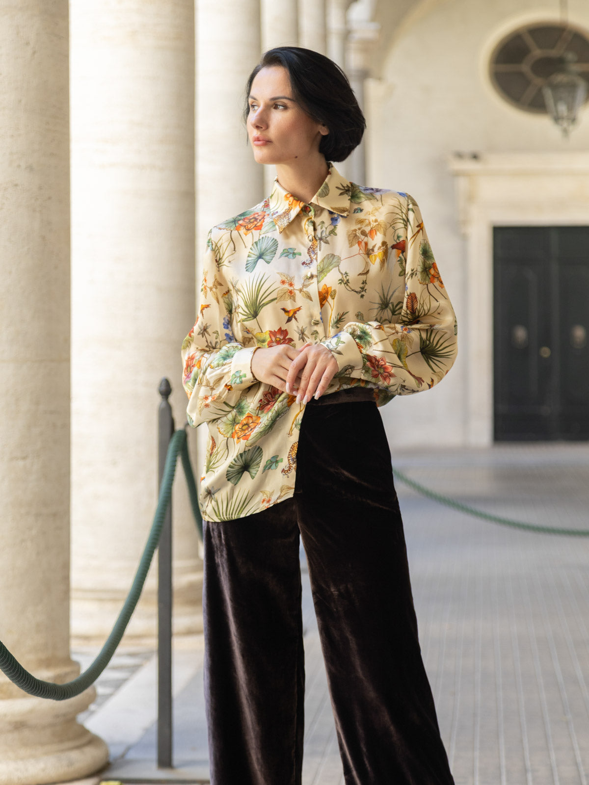 Modella in camicia floreale e pantalone velluto marrone palazzo, elegante e versatile in un ambiente raffinato.