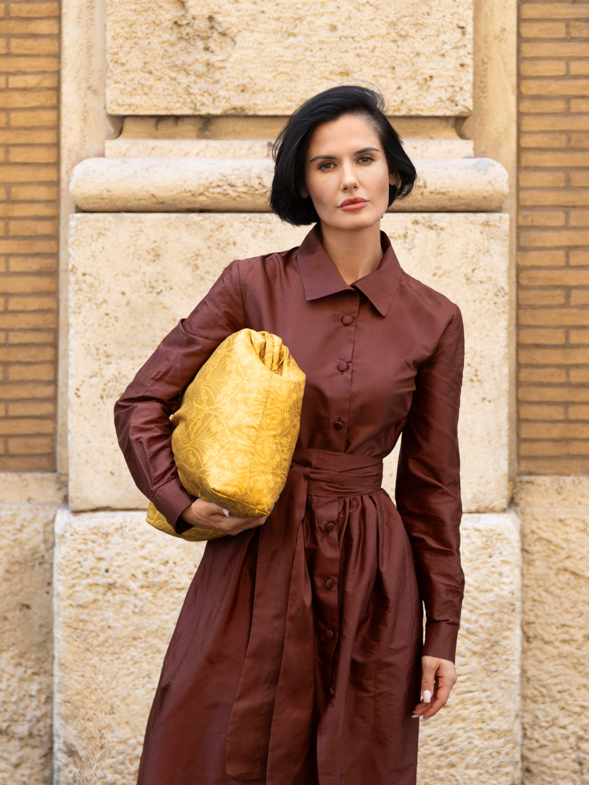 Modella in abito in shantung elegante marrone, con borsa gialla, davanti a muro di pietra.