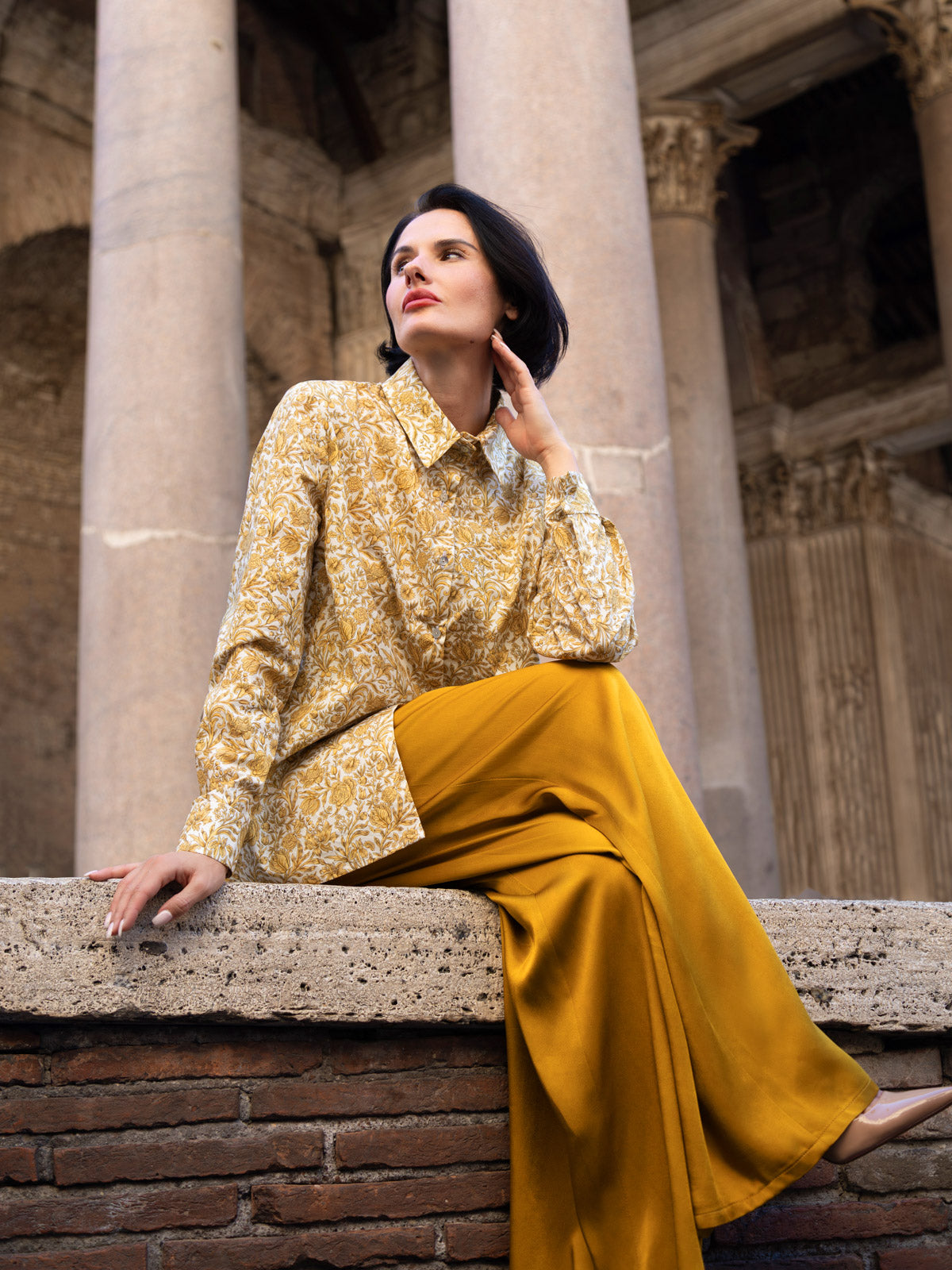 Modella che indossa una camicia Liberty in cotone con stampe eleganti, seduta su una pietra in un contesto architettonico.